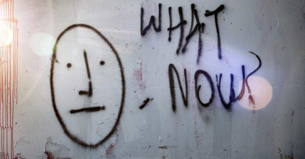 wall with chipping paint with a spray painting of a stick figure face and the words What Now?