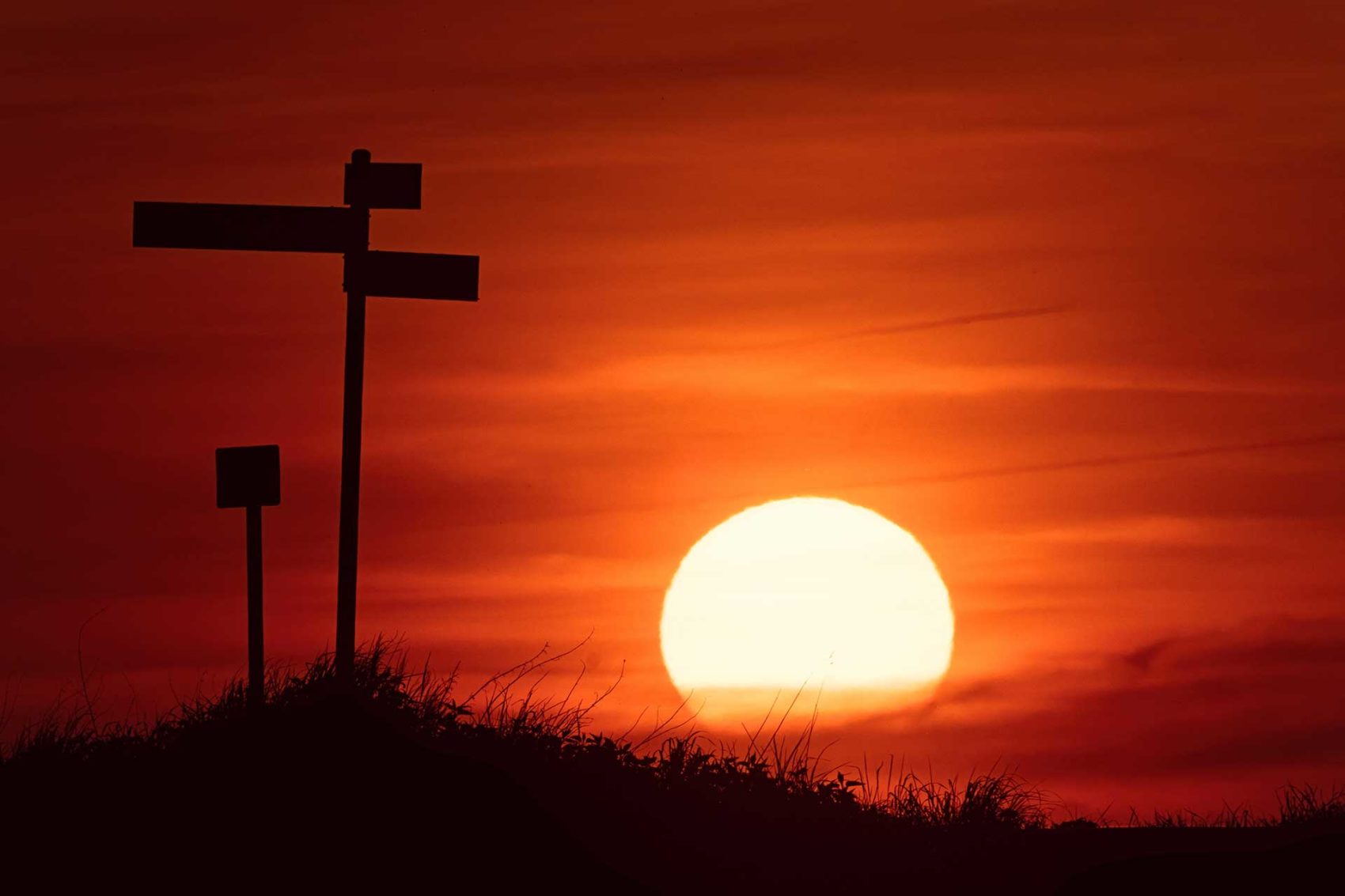 Roadside at dusk with a setting sun, orange sky, and a post with directional road signs